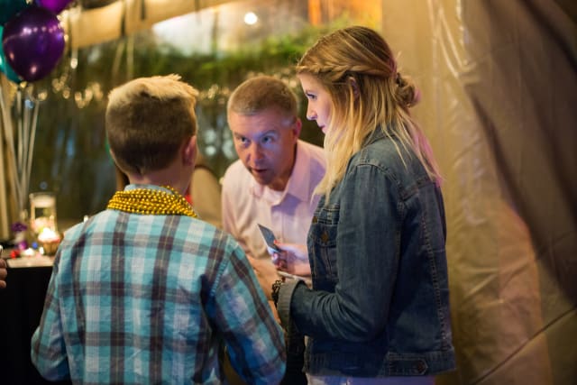 People engaging in conversation at the Love Bomb 2017 event, with a boy wearing a plaid shirt and beads, and a woman in a denim jacket, discussing with a man in a white shirt, set against a festive backdrop with balloons.